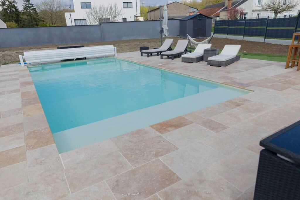 Piscine extérieure avec plage en dalles claires, margelles ton pierre, transats et jardin clôturé