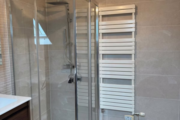 Salle d’eau rénovée avec douche d’angle vitrée, sèche-serviettes blanc, carrelage beige et sol imitation bois