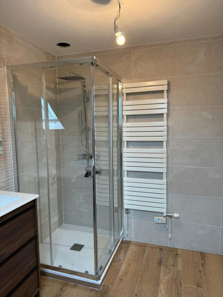 Salle d’eau rénovée avec douche d’angle vitrée, sèche-serviettes blanc, carrelage beige et sol imitation bois