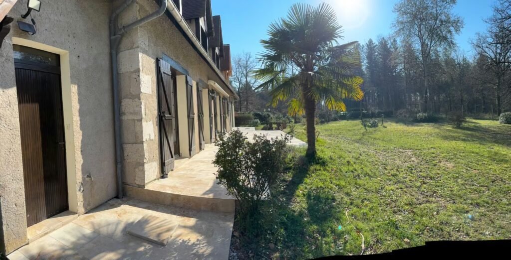 Terrasse extérieure rénovée en pierre claire le long d’une maison avec façade ancienne, jardin et pelouse