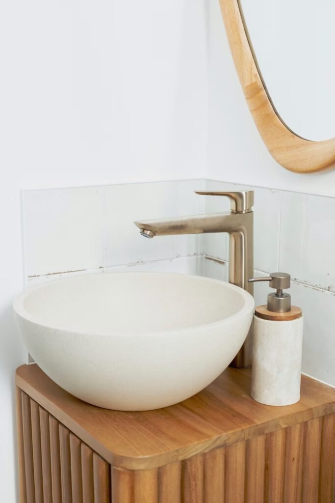 WC rénové avec lave-mains rond blanc, meuble bois à lattes, robinetterie murale dorée et miroir arrondi